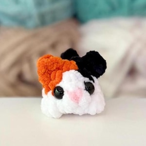 Puede incluir: Un peque&ntilde;o peluche de ganchillo redondo que se asemeja a un h&aacute;mster o cobaya. La criatura presenta un cuerpo blanco, una oreja naranja, una oreja negra, dos grandes ojos de pl&aacute;stico negro y una peque&ntilde;a nariz rosa bordada, hecho de lana.