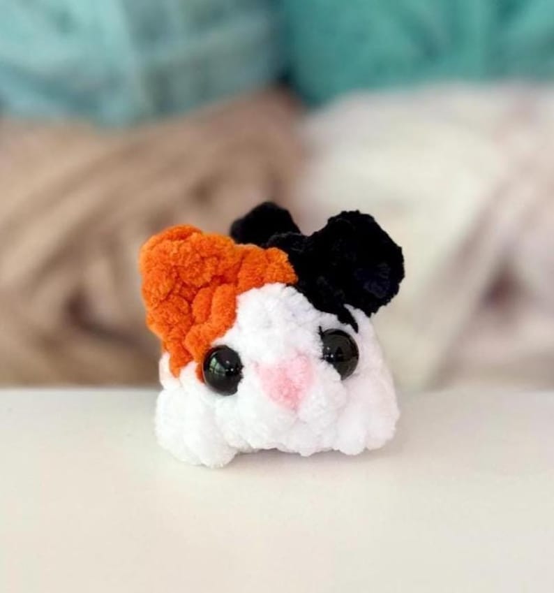 Puede incluir: Un peque&ntilde;o peluche de ganchillo redondo que se asemeja a un h&aacute;mster o cobaya. La criatura presenta un cuerpo blanco, una oreja naranja, una oreja negra, dos grandes ojos de pl&aacute;stico negro y una peque&ntilde;a nariz rosa bordada, hecho de lana.