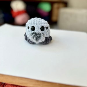 May include: A small gray crocheted plush toy with black eyes and a gray and black fuzzy nose. The toy is sitting on a white surface.