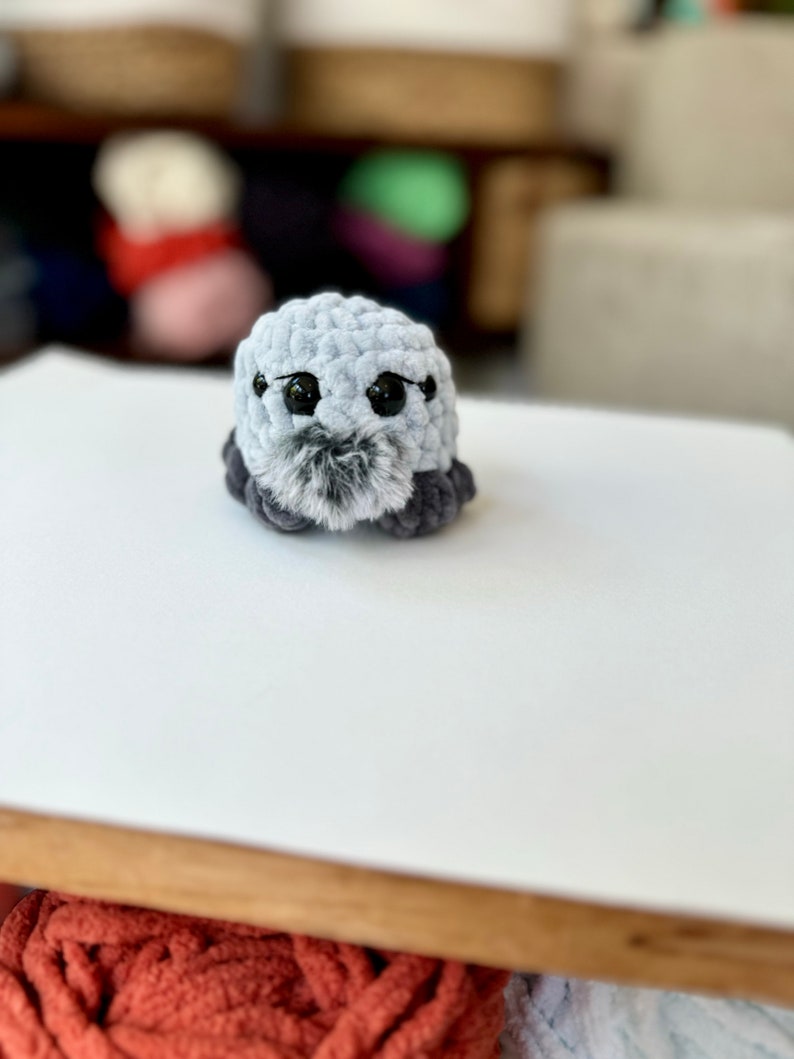 May include: A small gray crocheted plush toy with black eyes and a gray and black fuzzy nose. The toy is sitting on a white surface.