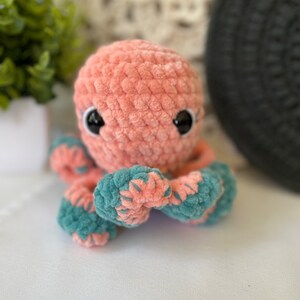Pode incluir: Um brinquedo de pel&uacute;cia de polvo de croch&ecirc; rosa e azul-petr&oacute;leo com olhos pretos.