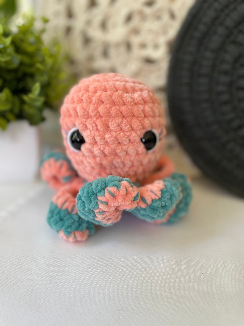 Pode incluir: Um brinquedo de pel&uacute;cia de polvo de croch&ecirc; rosa e azul-petr&oacute;leo com olhos pretos.