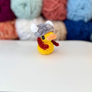 May include: A yellow crochet duck wearing a grey Viking helmet with white horns and a red scarf. The duck has an orange beak and black eyes. The background features colourful yarn balls.