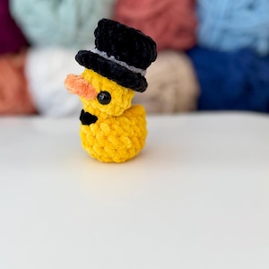 May include: A yellow crochet duck wearing a black top hat with a grey band and a black bow tie. The duck has an orange beak and black eyes. The background features colourful yarn.