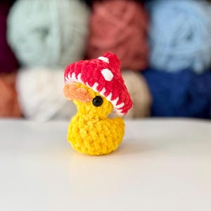 May include: A yellow crochet duck wearing a red mushroom hat with white spots. The duck has an orange beak and a black eye. The background features colourful yarn skeins.