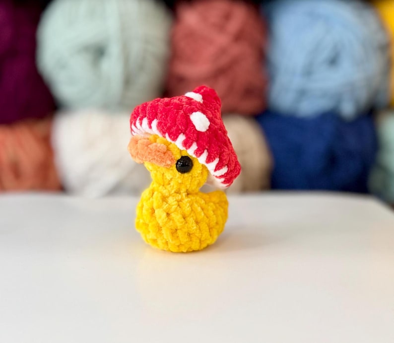 May include: A yellow crochet duck wearing a red mushroom hat with white spots. The duck has an orange beak and a black eye. The background features colourful yarn skeins.