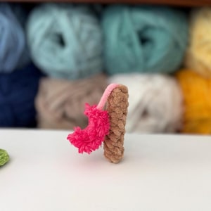 Puede incluir: Una peque&ntilde;a figura de ganchillo hecha a mano con forma de n&uacute;mero uno. El cuerpo principal est&aacute; elaborado con hilo marr&oacute;n claro con un acabado texturizado de bucles. Una pieza decorativa de color rosa brillante, difusa y texturizada est&aacute; unida al lateral con un tallo de hilo rosa fino.