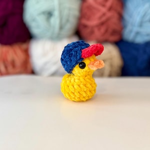 May include: A yellow crochet duck wearing a blue and red baseball cap. The duck has a small orange beak and black eyes. The background features various colours of yarn.