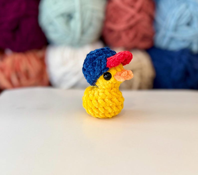 May include: A yellow crochet duck wearing a blue and red baseball cap. The duck has a small orange beak and black eyes. The background features various colours of yarn.
