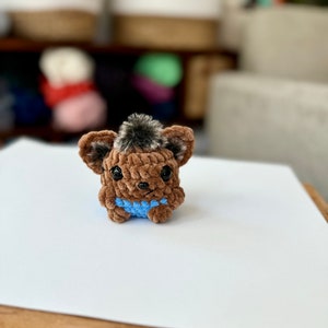 May include: A small brown crocheted animal with a blue crocheted vest. The animal has a fluffy brown tail and black eyes.