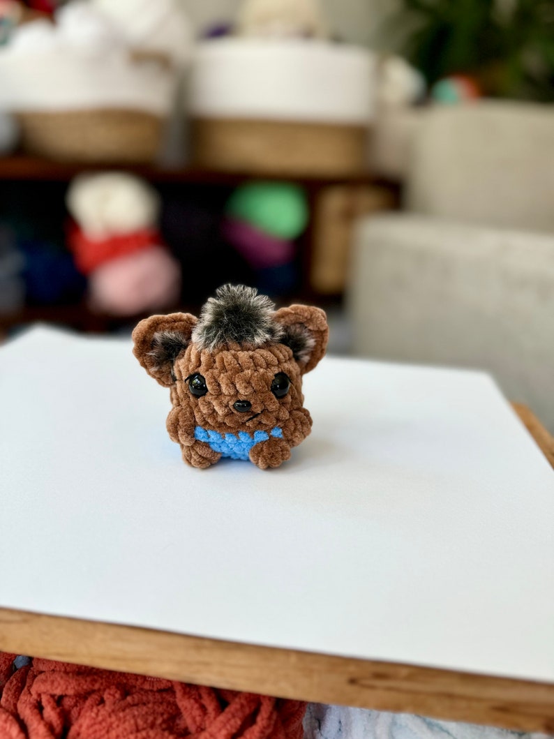 May include: A small brown crocheted animal with a blue crocheted vest. The animal has a fluffy brown tail and black eyes.