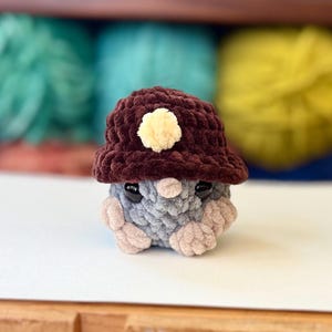 Op de afbeelding: Een klein gebreid pluche speelgoed dat een bruine gebreide hoed draagt met een gele pompon. Het speelgoed heeft grijze en roze gebreide details en zwarte ogen.