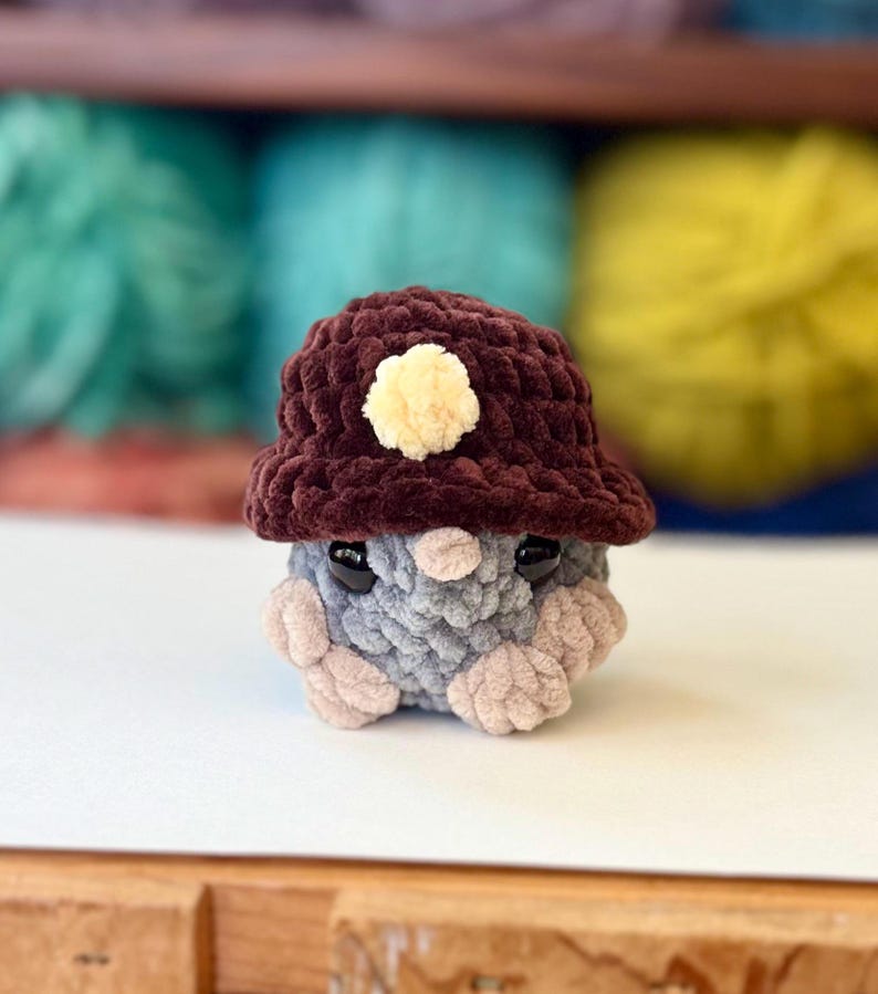 Op de afbeelding: Een klein gebreid pluche speelgoed dat een bruine gebreide hoed draagt met een gele pompon. Het speelgoed heeft grijze en roze gebreide details en zwarte ogen.