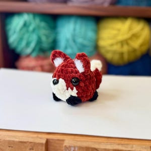 Op de afbeelding: Een klein, gehaakt knuffeldier in de vorm van een vos met een rood en wit lichaam, zwarte ogen en een zwarte neus. De vos zit op een wit oppervlak.
