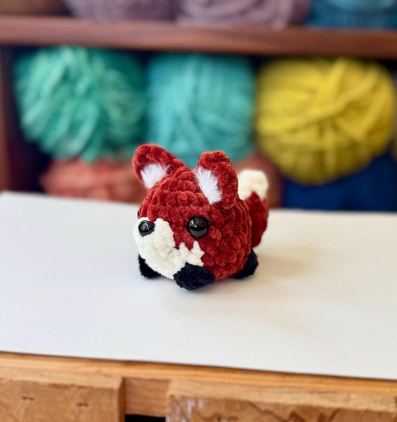 Op de afbeelding: Een klein, gehaakt knuffeldier in de vorm van een vos met een rood en wit lichaam, zwarte ogen en een zwarte neus. De vos zit op een wit oppervlak.
