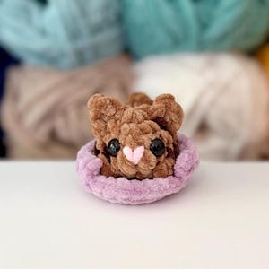 Puede incluir: Un peque&ntilde;o animal de peluche tejido a ganchillo en color marr&oacute;n con grandes ojos negros y una nariz rosa claro en forma de coraz&oacute;n. La figura est&aacute; colocada dentro de una base circular de ganchillo color violeta claro, suave y texturizada.
