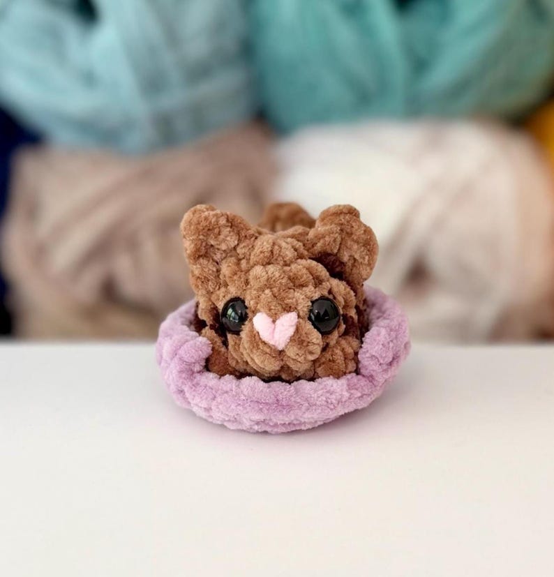 Puede incluir: Un peque&ntilde;o animal de peluche tejido a ganchillo en color marr&oacute;n con grandes ojos negros y una nariz rosa claro en forma de coraz&oacute;n. La figura est&aacute; colocada dentro de una base circular de ganchillo color violeta claro, suave y texturizada.