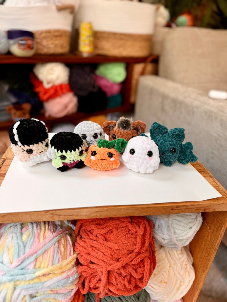 May include: A collection of eight crocheted Halloween-themed characters, including a ghost, a pumpkin, a monster, and a vampire. The characters are made with different colored yarn and have various facial expressions.