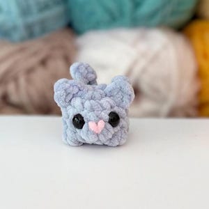 Puede incluir: Un peque&ntilde;o peluche de ganchillo de color azul claro con forma de gato. La figura presenta una superficie de hilo suave y texturizada, dos orejas redondeadas, una peque&ntilde;a cola, dos grandes ojos de seguridad de pl&aacute;stico negro y una peque&ntilde;a nariz cosida en forma de coraz&oacute;n de color rosa claro.