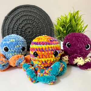 Pode incluir: Tr&ecirc;s peluches de polvo de croch&ecirc; em diferentes cores. Um &eacute; azul com tent&aacute;culos laranja, outro &eacute; roxo com tent&aacute;culos amarelos e o &uacute;ltimo &eacute; uma mistura de amarelo, laranja, rosa e azul com tent&aacute;culos turquesa.