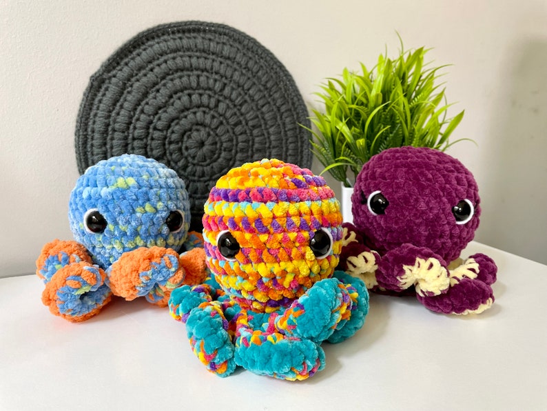 Pode incluir: Tr&ecirc;s peluches de polvo de croch&ecirc; em diferentes cores. Um &eacute; azul com tent&aacute;culos laranja, outro &eacute; roxo com tent&aacute;culos amarelos e o &uacute;ltimo &eacute; uma mistura de amarelo, laranja, rosa e azul com tent&aacute;culos turquesa.