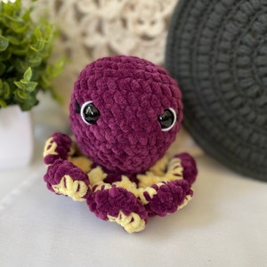Pode incluir: Um polvo de croch&ecirc; roxo com detalhes amarelos e olhos pretos. O polvo est&aacute; sentado em uma superf&iacute;cie branca.