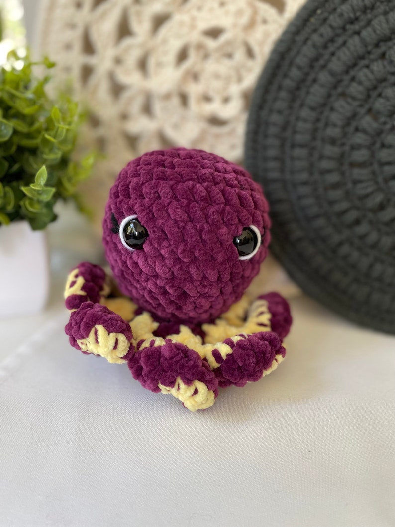 Pode incluir: Um polvo de croch&ecirc; roxo com detalhes amarelos e olhos pretos. O polvo est&aacute; sentado em uma superf&iacute;cie branca.