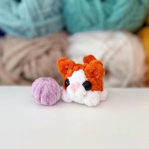 Puede incluir: Un peque&ntilde;o gato de peluche tejido a ganchillo con hilo naranja y blanco, con ojos de pl&aacute;stico negro y nariz rosa, colocado junto a una diminuta bola de hilo redonda tejida a ganchillo de color violeta claro. El gato tiene textura suave.