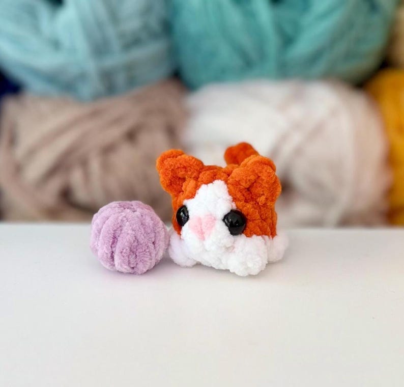 Puede incluir: Un peque&ntilde;o gato de peluche tejido a ganchillo con hilo naranja y blanco, con ojos de pl&aacute;stico negro y nariz rosa, colocado junto a una diminuta bola de hilo redonda tejida a ganchillo de color violeta claro. El gato tiene textura suave.