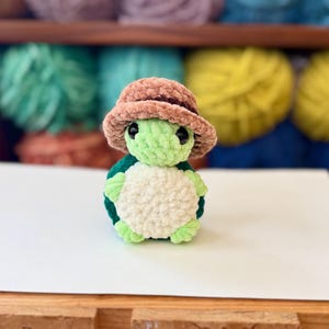 Op de afbeelding: Een kleine groene gehaakte schildpad met een bruine gehaakte hoed. De schildpad zit op een wit oppervlak.