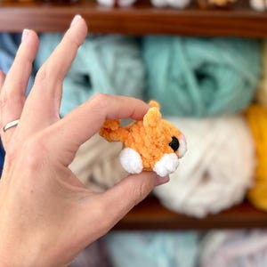 Puede incluir: Una peque&ntilde;a criatura de hilo naranja y blanca hecha a mano con una textura difusa, que presenta un cuerpo redondeado, una cola peque&ntilde;a y ojos de pl&aacute;stico negro prominentes. La figura en miniatura est&aacute; hecha de fibras de hilo suaves y afelpadas.