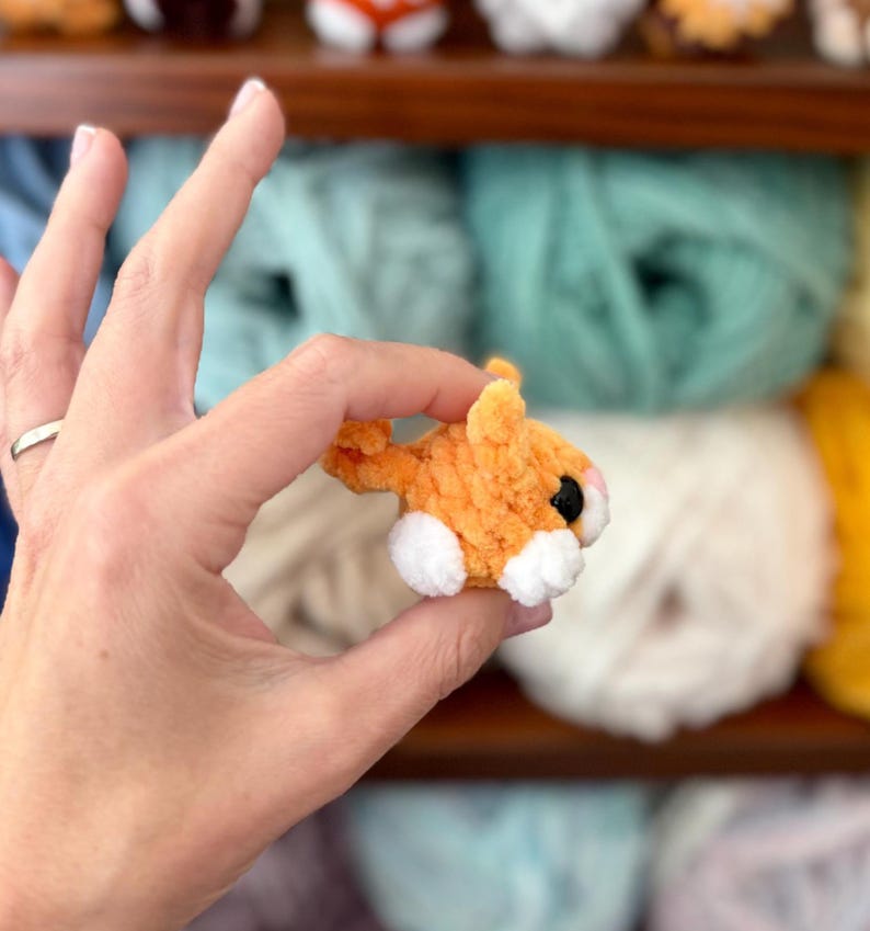 Puede incluir: Una peque&ntilde;a criatura de hilo naranja y blanca hecha a mano con una textura difusa, que presenta un cuerpo redondeado, una cola peque&ntilde;a y ojos de pl&aacute;stico negro prominentes. La figura en miniatura est&aacute; hecha de fibras de hilo suaves y afelpadas.