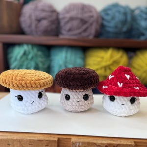 Op de afbeelding: Drie gehaakte pluche paddenstoelen met verschillende gekleurde hoeden. De eerste paddenstoel heeft een gele hoed, de tweede een bruine hoed en de derde een rode hoed. Alle drie de paddenstoelen hebben witte lichamen en schattige gezichten.