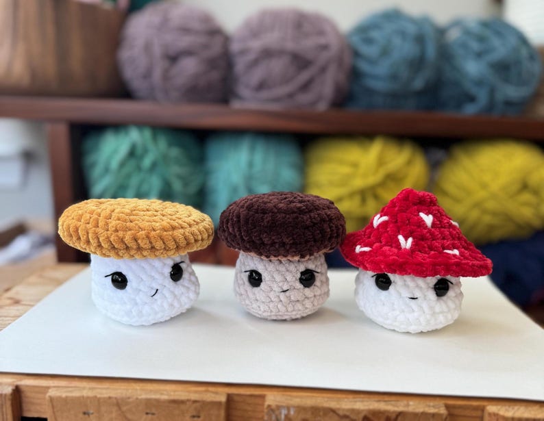 Op de afbeelding: Drie gehaakte pluche paddenstoelen met verschillende gekleurde hoeden. De eerste paddenstoel heeft een gele hoed, de tweede een bruine hoed en de derde een rode hoed. Alle drie de paddenstoelen hebben witte lichamen en schattige gezichten.