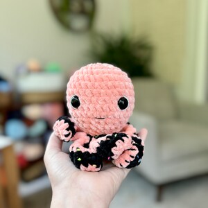 Pode incluir: Um polvo de croch&ecirc; rosa com tent&aacute;culos listrados de preto e branco. O polvo tem olhos grandes e pretos e um pequeno sorriso rosa.