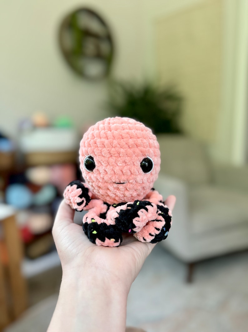 Pode incluir: Um polvo de croch&ecirc; rosa com tent&aacute;culos listrados de preto e branco. O polvo tem olhos grandes e pretos e um pequeno sorriso rosa.