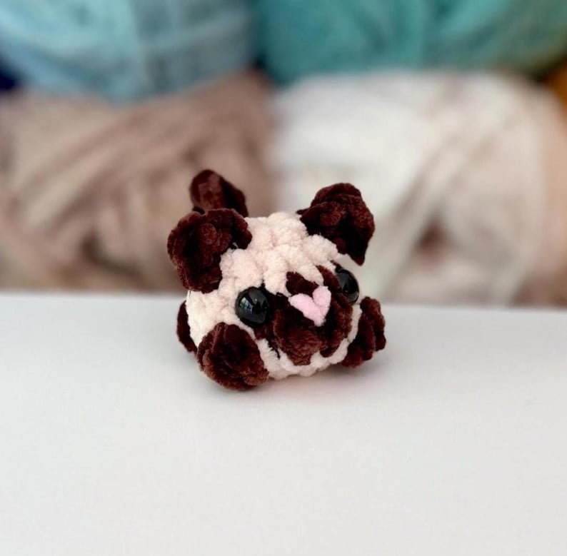 Puede incluir: Un peque&ntilde;o juguete de peluche tejido a ganchillo con forma de perro pug. La figura presenta un cuerpo de color crema con manchas de color marr&oacute;n oscuro alrededor de las orejas, los ojos y el hocico. Tiene dos ojos grandes, redondos y negros de pl&aacute;stico y una peque&ntilde;a nariz triangular de color rosa claro.