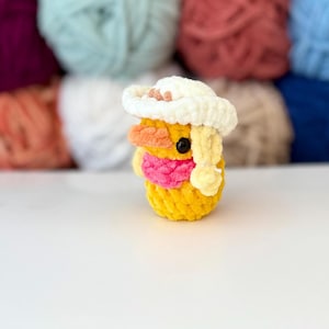 May include: A handmade yellow crochet duck wearing a white cowboy hat with a pink scarf. The duck has orange and yellow accents and a black eye. Balls of colourful yarn are in the background.