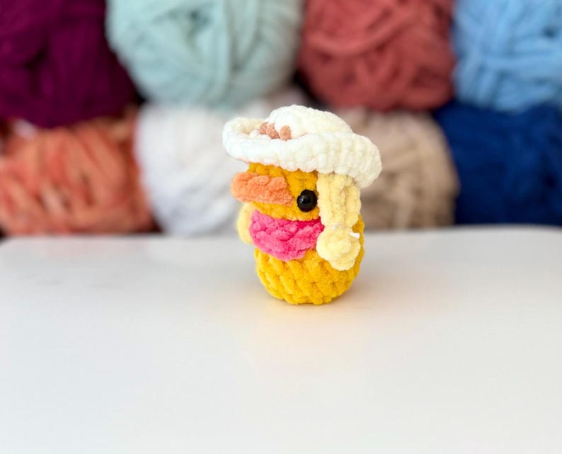 May include: A handmade yellow crochet duck wearing a white cowboy hat with a pink scarf. The duck has orange and yellow accents and a black eye. Balls of colourful yarn are in the background.