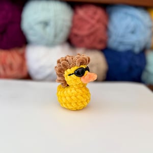 May include: A yellow crochet duck with black sunglasses and a brown wig. The duck has an orange beak and is made of textured yarn. The background shows various coloured yarn skeins, suitable for crafting.