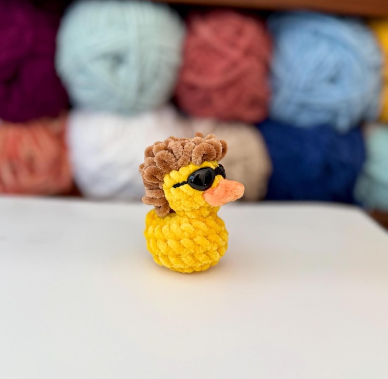 May include: A yellow crochet duck with black sunglasses and a brown wig. The duck has an orange beak and is made of textured yarn. The background shows various coloured yarn skeins, suitable for crafting.