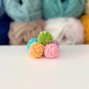 Puede incluir: Un grupo de cinco pompones peque&ntilde;os, redondos y difusos hechos de hilo suave. Los pompones est&aacute;n dispuestos en un peque&ntilde;o mont&oacute;n y presentan colores vibrantes que incluyen verde lima, naranja brillante, rosa claro, azul turquesa y lavanda.
