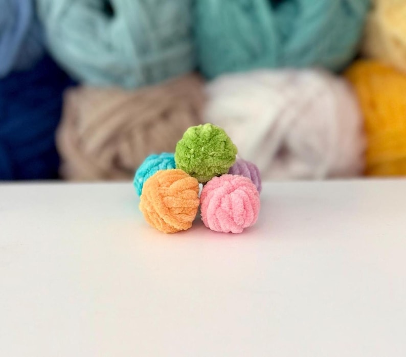 Puede incluir: Un grupo de cinco pompones peque&ntilde;os, redondos y difusos hechos de hilo suave. Los pompones est&aacute;n dispuestos en un peque&ntilde;o mont&oacute;n y presentan colores vibrantes que incluyen verde lima, naranja brillante, rosa claro, azul turquesa y lavanda.