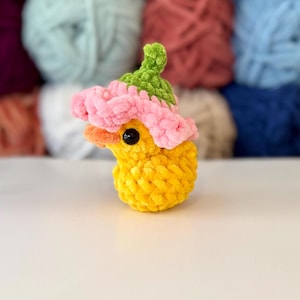 May include: A handmade yellow crochet duck wearing a pink and green flower hat. The duck has a small orange beak and black eyes. The background features several balls of yarn in various colours.