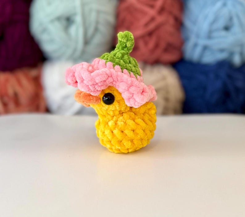 May include: A handmade yellow crochet duck wearing a pink and green flower hat. The duck has a small orange beak and black eyes. The background features several balls of yarn in various colours.
