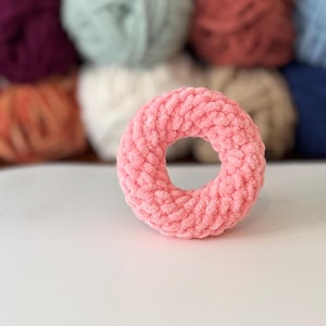May include: A light pink, donut-shaped crochet item. The yarn has a soft, textured appearance. The background features several balls of yarn in various colours, including purple, green, and brown.