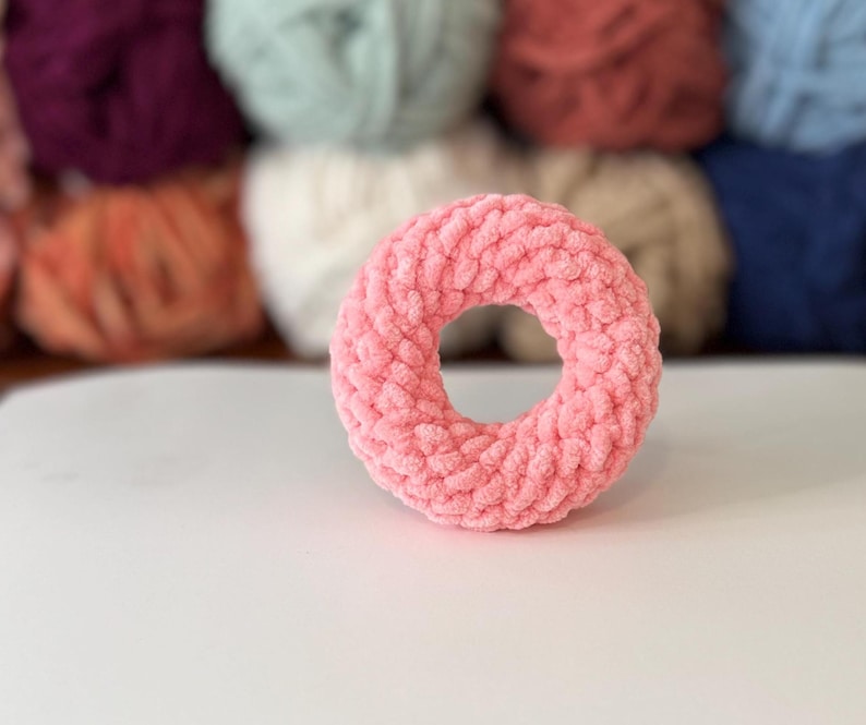 May include: A light pink, donut-shaped crochet item. The yarn has a soft, textured appearance. The background features several balls of yarn in various colours, including purple, green, and brown.