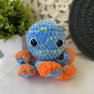 Pode incluir: Um polvo de croch&ecirc; azul e laranja com detalhes verdes. O polvo tem olhos pretos grandes e uma textura macia e felpuda.