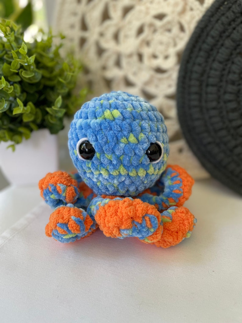 Pode incluir: Um polvo de croch&ecirc; azul e laranja com detalhes verdes. O polvo tem olhos pretos grandes e uma textura macia e felpuda.