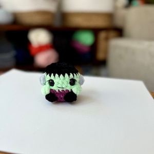 May include: A small green crocheted monster with black hair, purple clothing, and big black eyes. The monster is sitting on a white surface.